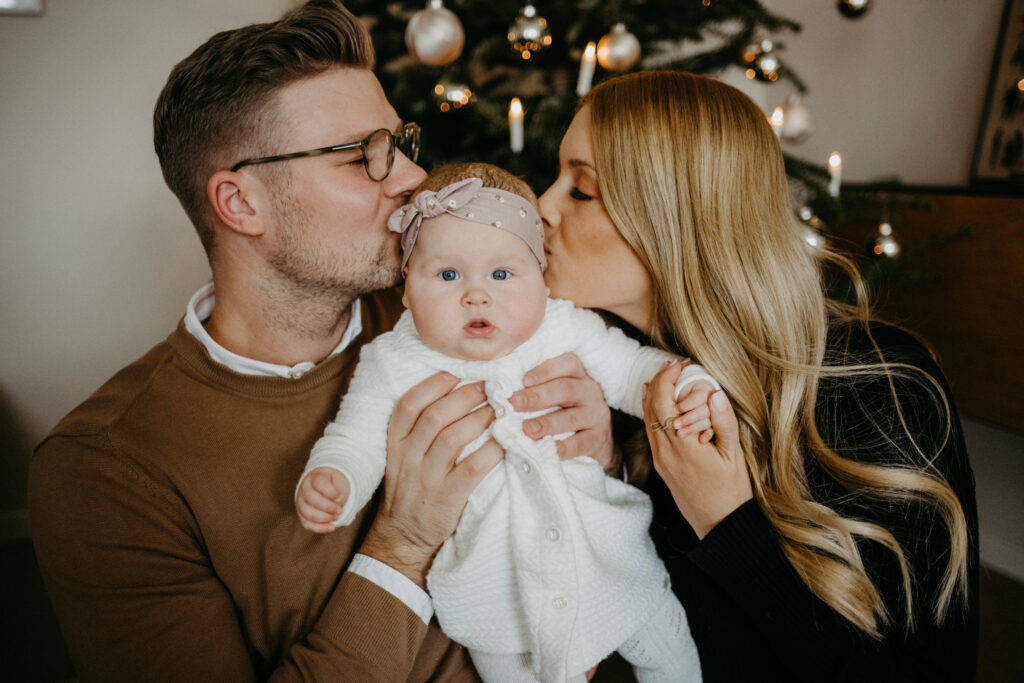 Eltern küssen ihr baby vor dem Weihnachtsbaum beim Weihnachtsmarkt-Fotoshooting für Familien in Bonn