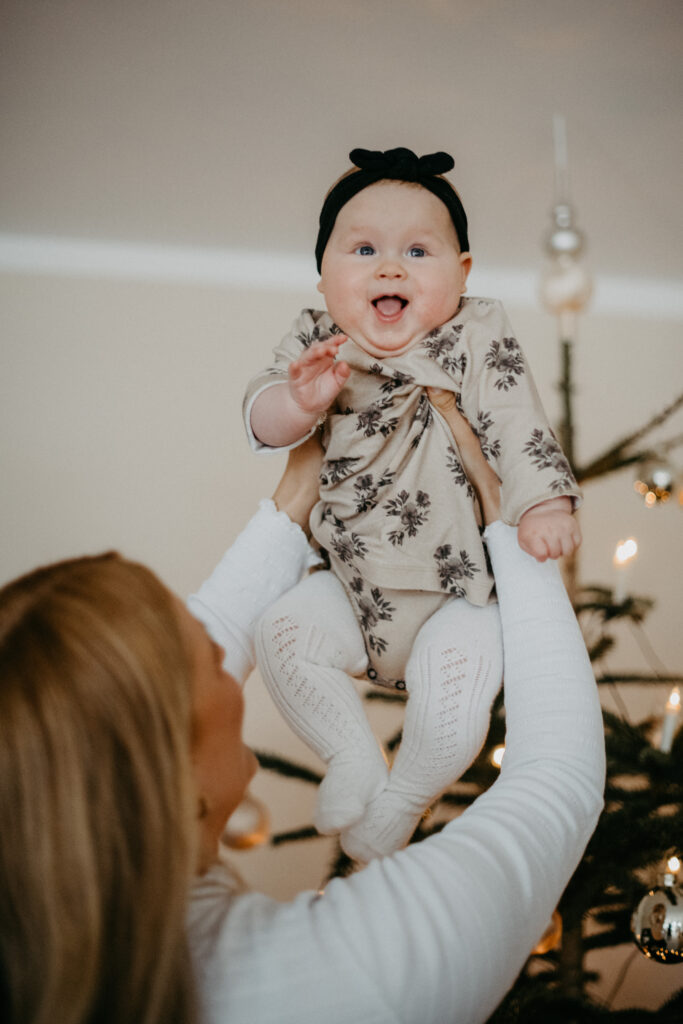 Baby freut sich sehr, als die Mutter es vor dem Weihnachtsbaum in die Höhe hält.