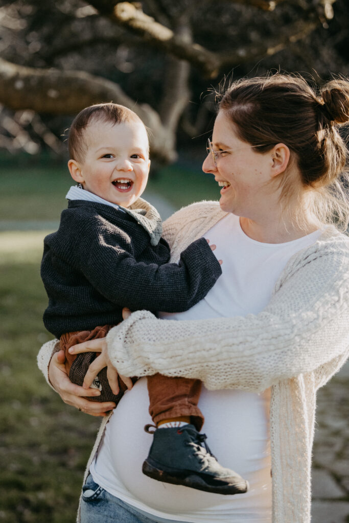Eine Schwangere hält beim Schwangerschafts-Fotoshooting ihren älteren Sohn auf dem Arm, der lacht. Es ist Winter.