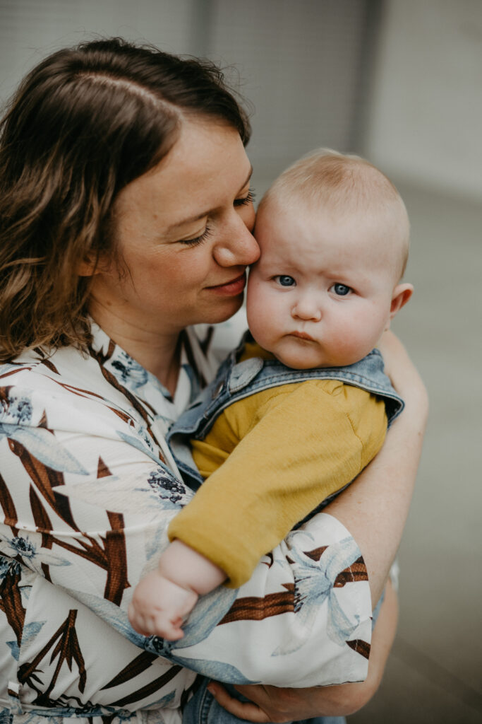 Babyfotoshooting in Bonn. Die Mutter schmiegt sich mit ihrem Gesicht ans baby, das neugierig die Fotografin beobachtet